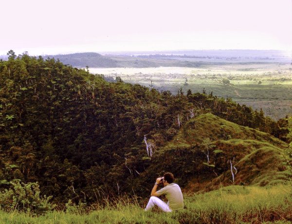 Man sits in field scanning forested hill with binoculars, looking for birds.