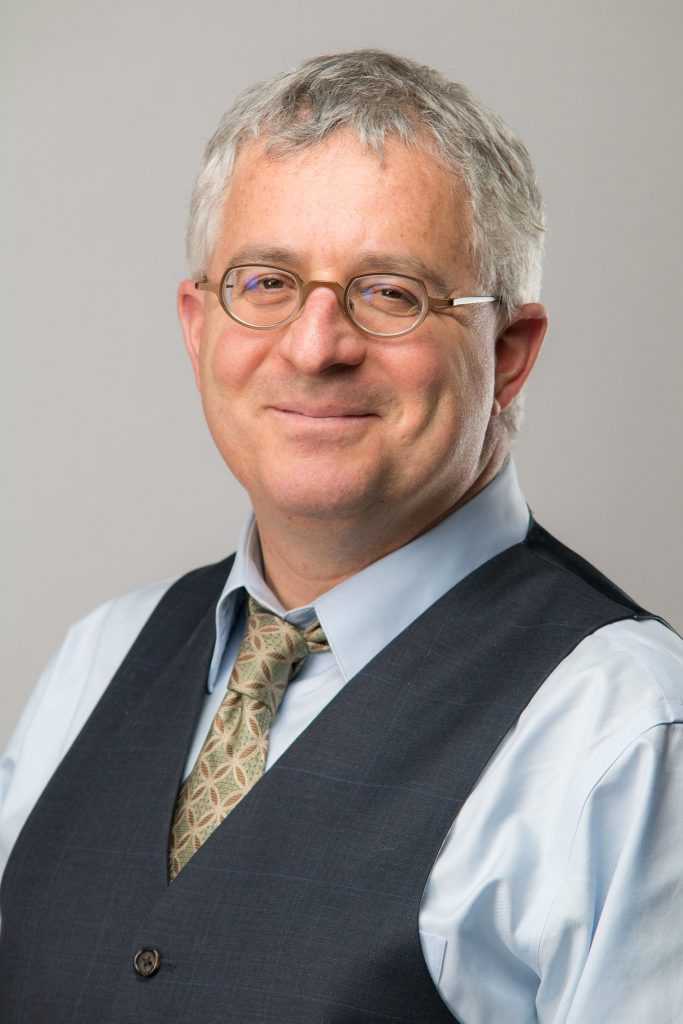 Portrait of man with small round glasses and grey hair wearing shirt, tie and vest. Man is looking directly at camera and smiling.