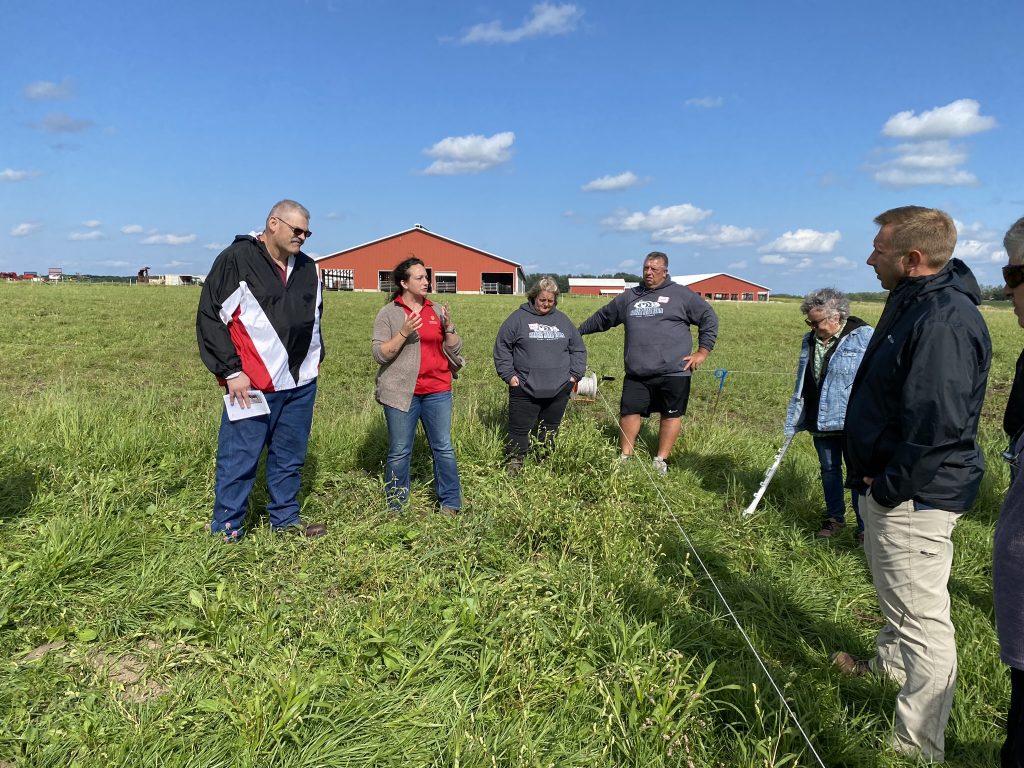 A group of six people standing in a grassy field during an outdoor discussion. One person in the center appears to be speaking to the group. In the background, there are red barns and a blue sky.