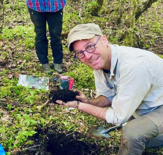 Man in hat and glasses kneeling in field with a soil sample in his hands.