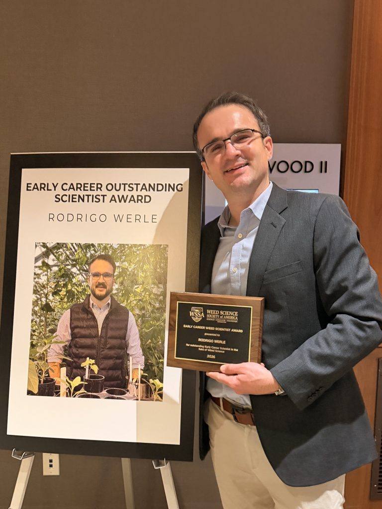 Man in glasses, blue shirt, gray jacket and khaki pants stands with plaque.