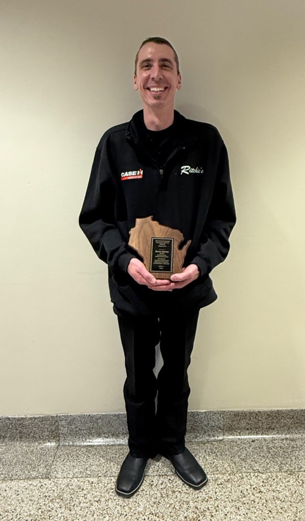 Man in black pants and shirt standing with plaque in the shape of Wisconsin.