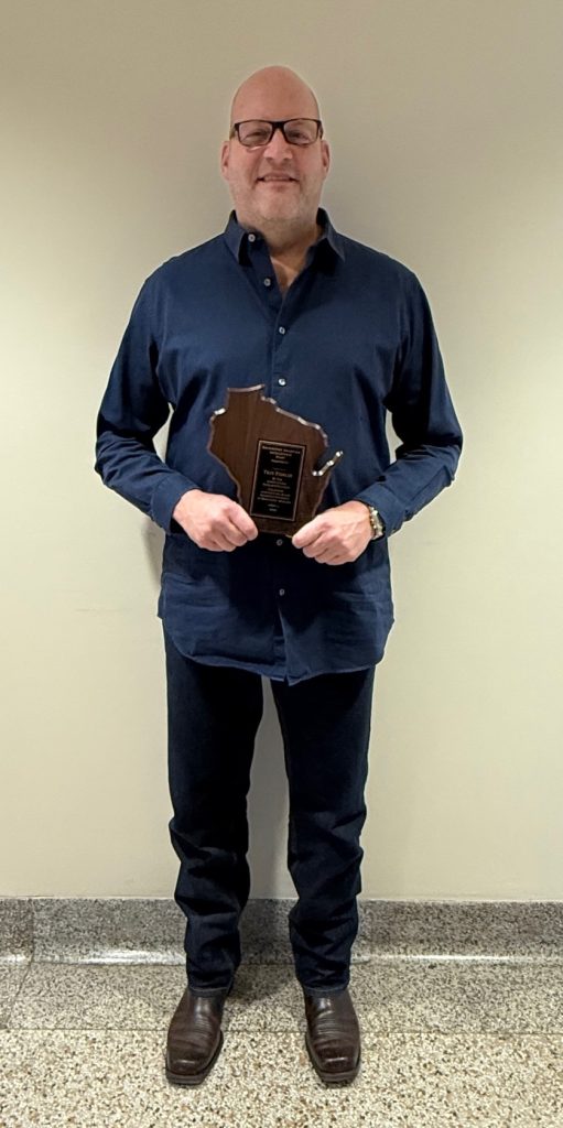 Man in blue shirt and black pants standing with plaque in shape of Wisconsin.