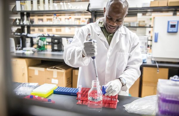 Smiling man in lab coat stands at bench with lab equipment in front of him.