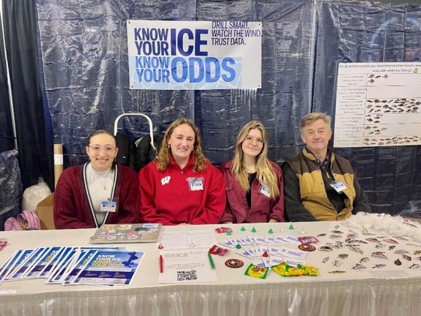 Three women and one man sit behind table covered with flyers and giveaways.