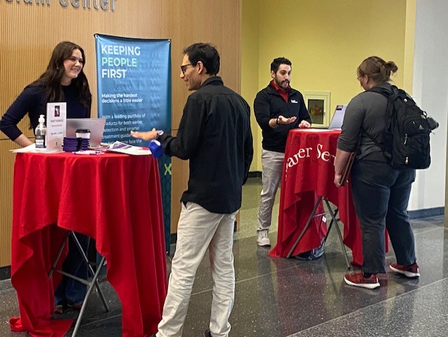 Two tables with red tableclothes in a lobby with two people at each table talking to each other.