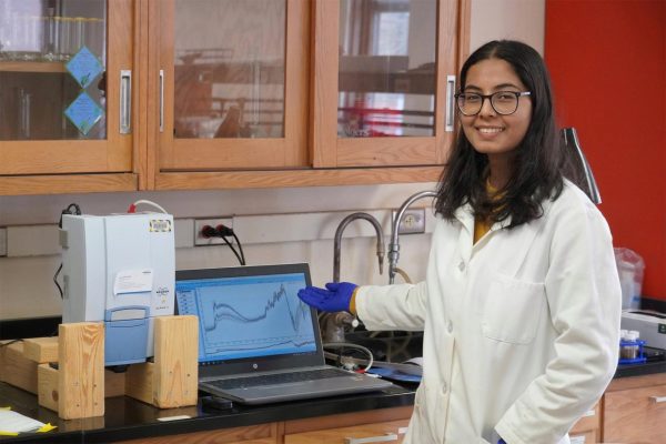Woman scientist in lab coat and glasses stands in front of lab bench showing a graph on a computer.