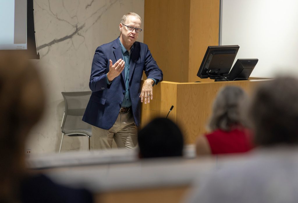 Man in blue jacket stands leaning on podium at front of room talking to group