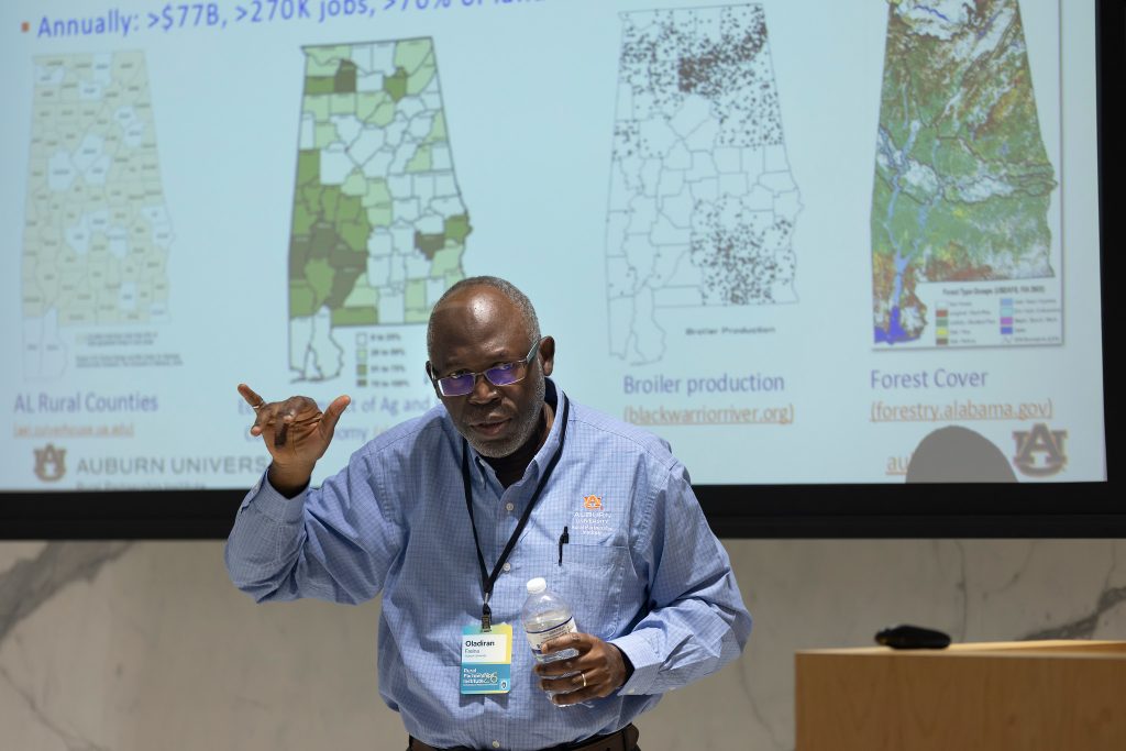 Man in blue shirt and glasses stands in front of maps on a screen talking