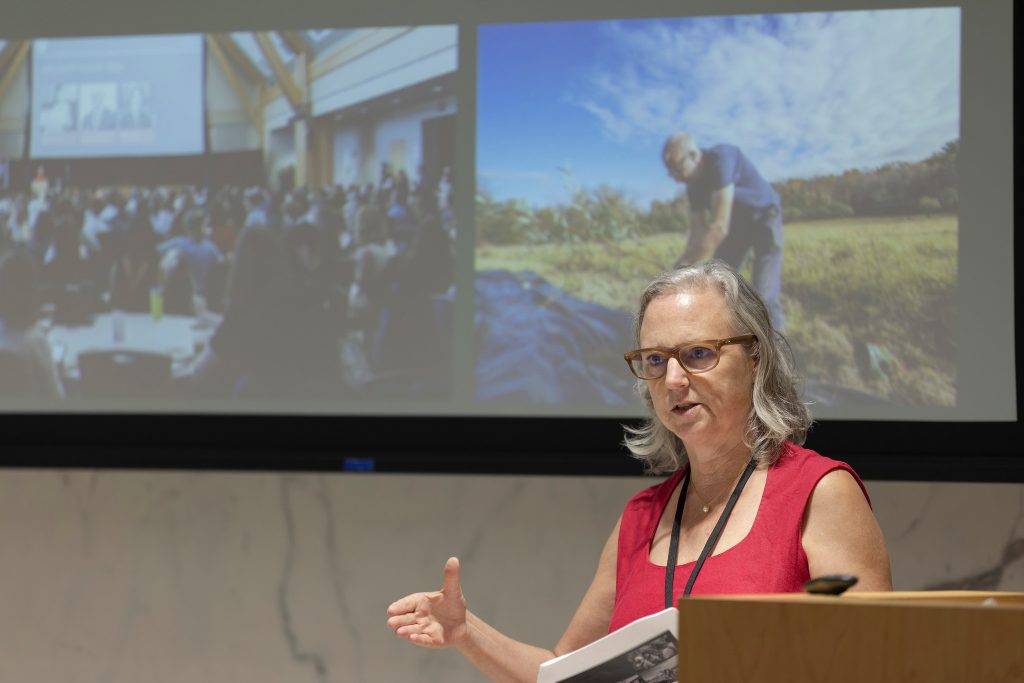 Woman in red dress and glasses stands in front of photos on large screen talking