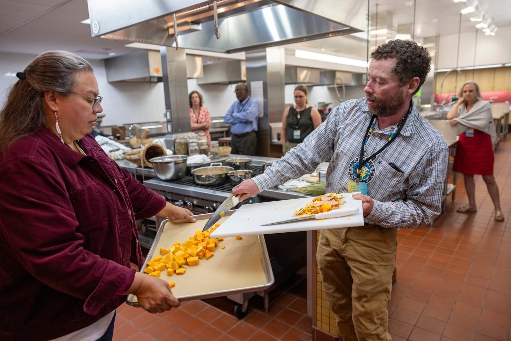 Woman and man transfer squash from a cutting board to a large baking sheet while others look on