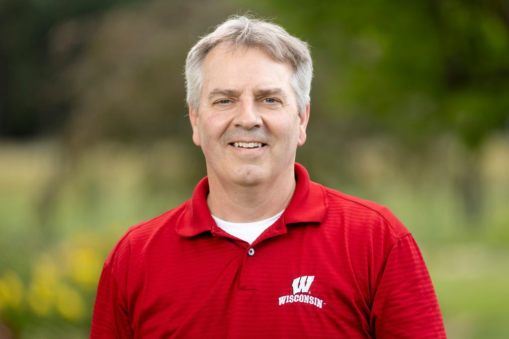 Man with grey hair and red Wisconsin shirt smiles at camera.