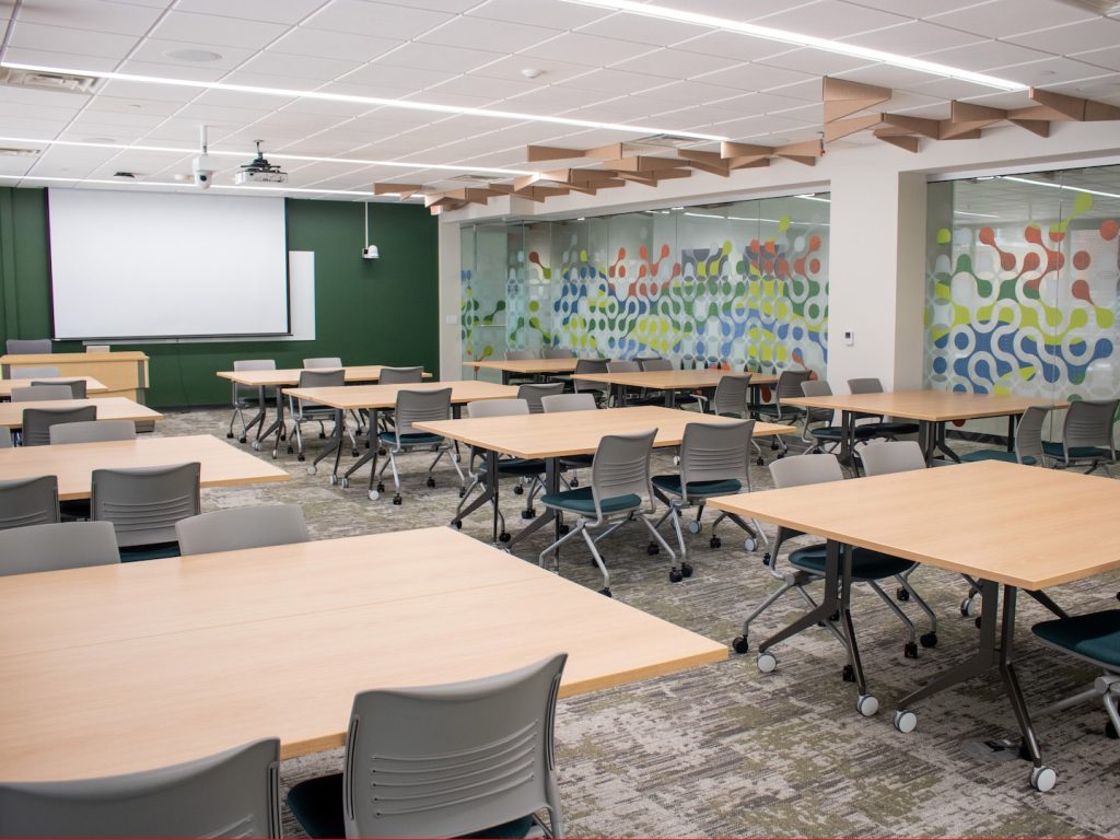 Rows of tables with chairs fill a large room with grey carpet and colorful designs on one wall. On the far wall is a projector screen.