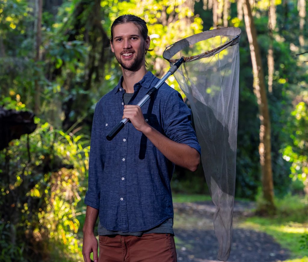 A smiling man holding a large insect net in a forest setting.